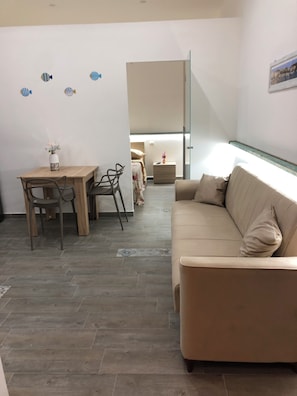 A modern living room with a beige sofa, a wooden dining table with chairs, and a small table with a vase. - Michelina Vacation Home: An apartment of about 45 m² equipped with everything you need (Monte di Procida)