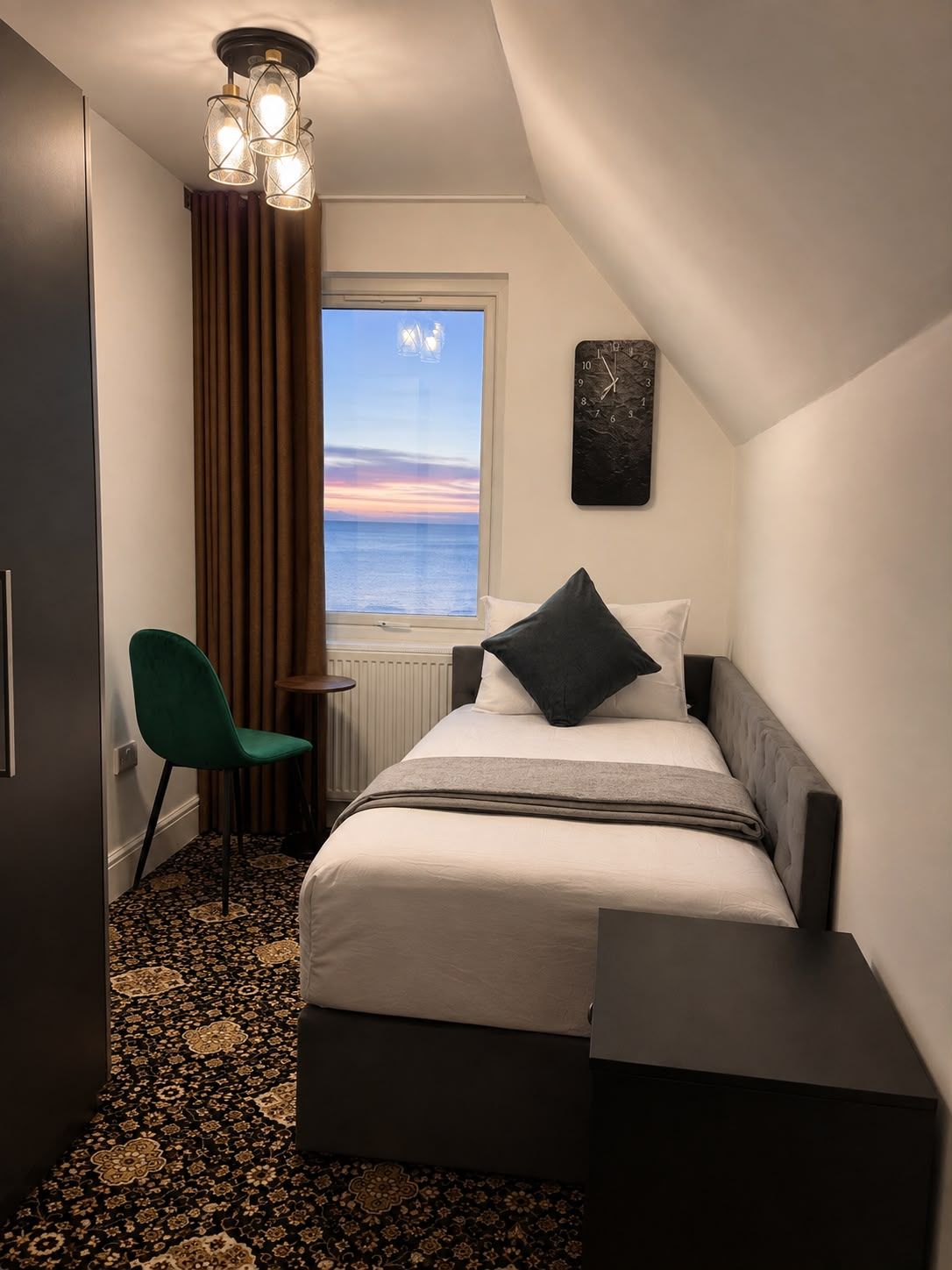 A hotel room with a bed, a chair, a window with a view of the sea, and a clock on the wall.