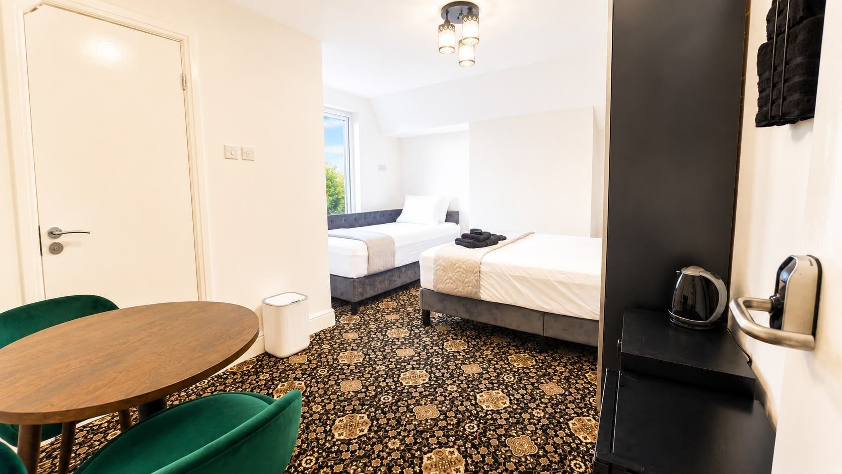A hotel room with a bed, a table, and a chair. There is a window with a view of greenery outside.