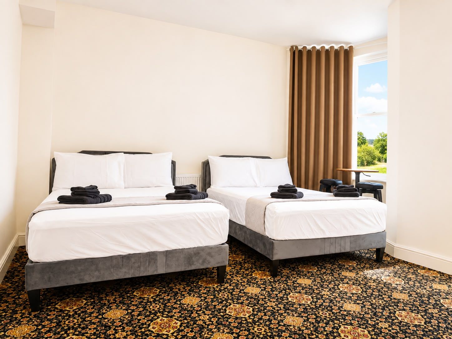 A hotel room with two beds, a window with curtains, a small table, and a patterned carpet.