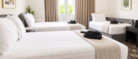 Quarto de hotel com duas camas, janela com cortinas, mesa de cabeceira com chaleira e planta emoldurada na parede.