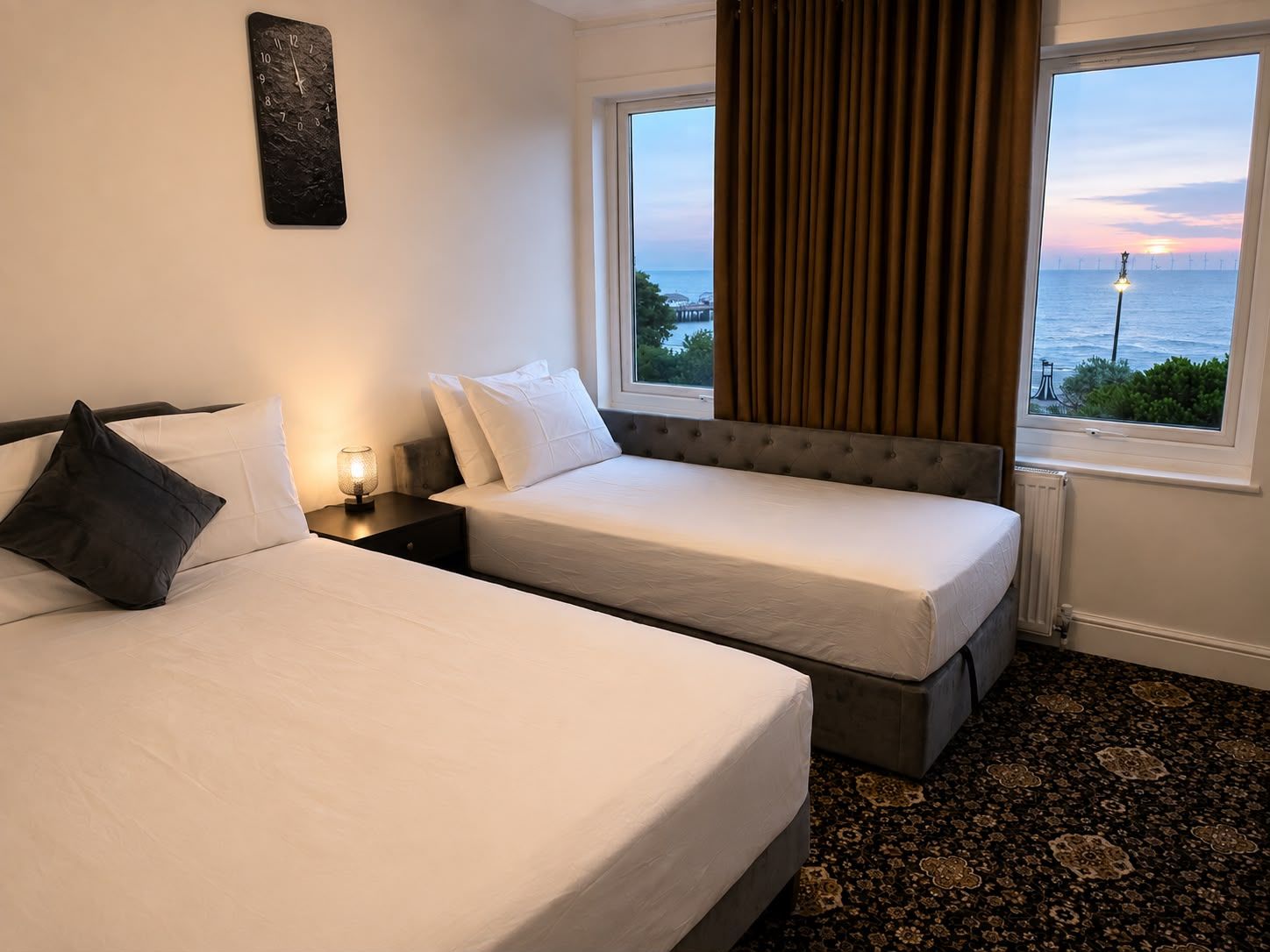 A hotel room with two beds, a nightstand, a lamp, a view of the sea, and a large window with curtains.