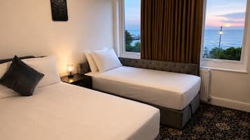 A hotel room with two beds, a nightstand, a lamp, a view of the sea, and a large window with curtains.