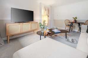 A modern living room with a flat-screen TV, a wooden cabinet, a round coffee table, a sofa, and a dining area with chairs and a small table. - Modern 1-bed-Parking-Garden-Nr Warner Bros (Watford)