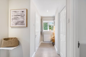 A hallway with white walls, a framed abstract painting, a woven basket on a table, and a window with a view of greenery. - Modern 1-bed-Parking-Garden-Nr Warner Bros (Watford)