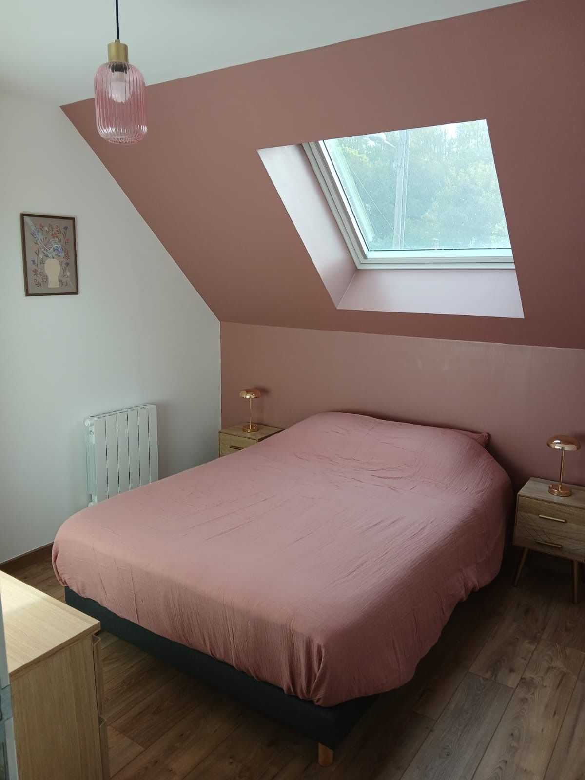 Un dormitorio con techo inclinado, tragaluces, cama con colcha rosa, mesita de noche con lámpara y radiador.