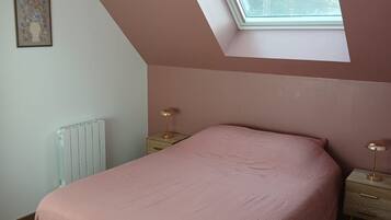 Un dormitorio con techo inclinado, tragaluces, cama con colcha rosa, mesita de noche con lámpara y radiador.