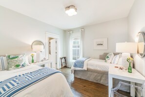 Quarto de hotel com duas camas, uma escrivaninha, uma cadeira, um espelho, uma janela com cortinas e um quadro na parede.