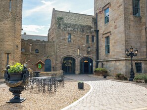 Ein Innenhof mit einem Steinhaus, einem Schild mit der Aufschrift 'Welcome to the Castle', einem schmiedeeisernen Tisch und Stühlen sowie einem Kopfsteinpflasterweg.