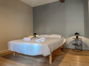 Een keurig opgemaakt bed met witte lakens en handdoeken, een nachtkastje met een lamp, en een klein rond tafeltje met een lamp.