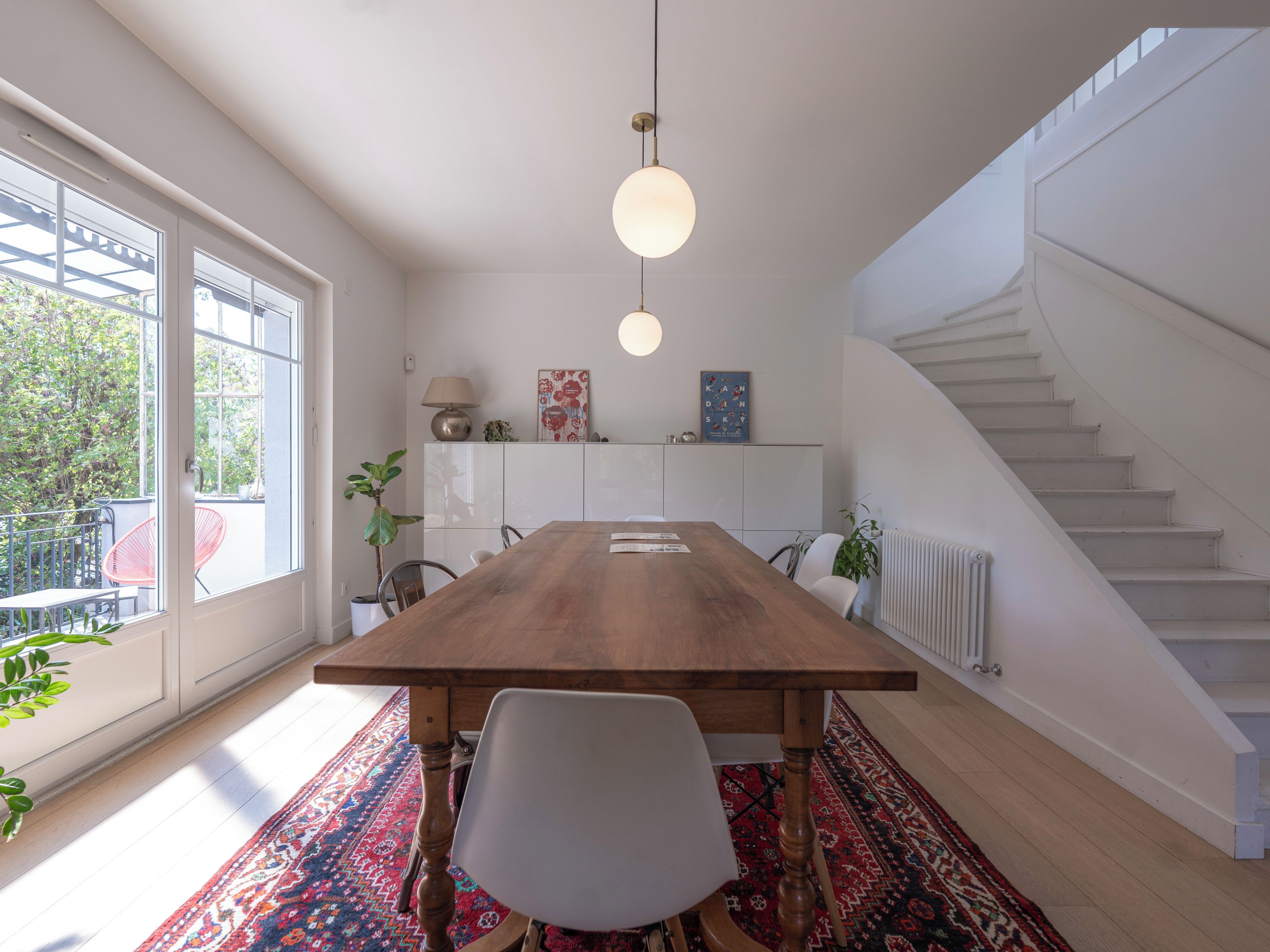 Un espace de salle à manger moderne avec une table en bois, des chaises blanches, un luminaire suspendu et une vue sur un escalier.