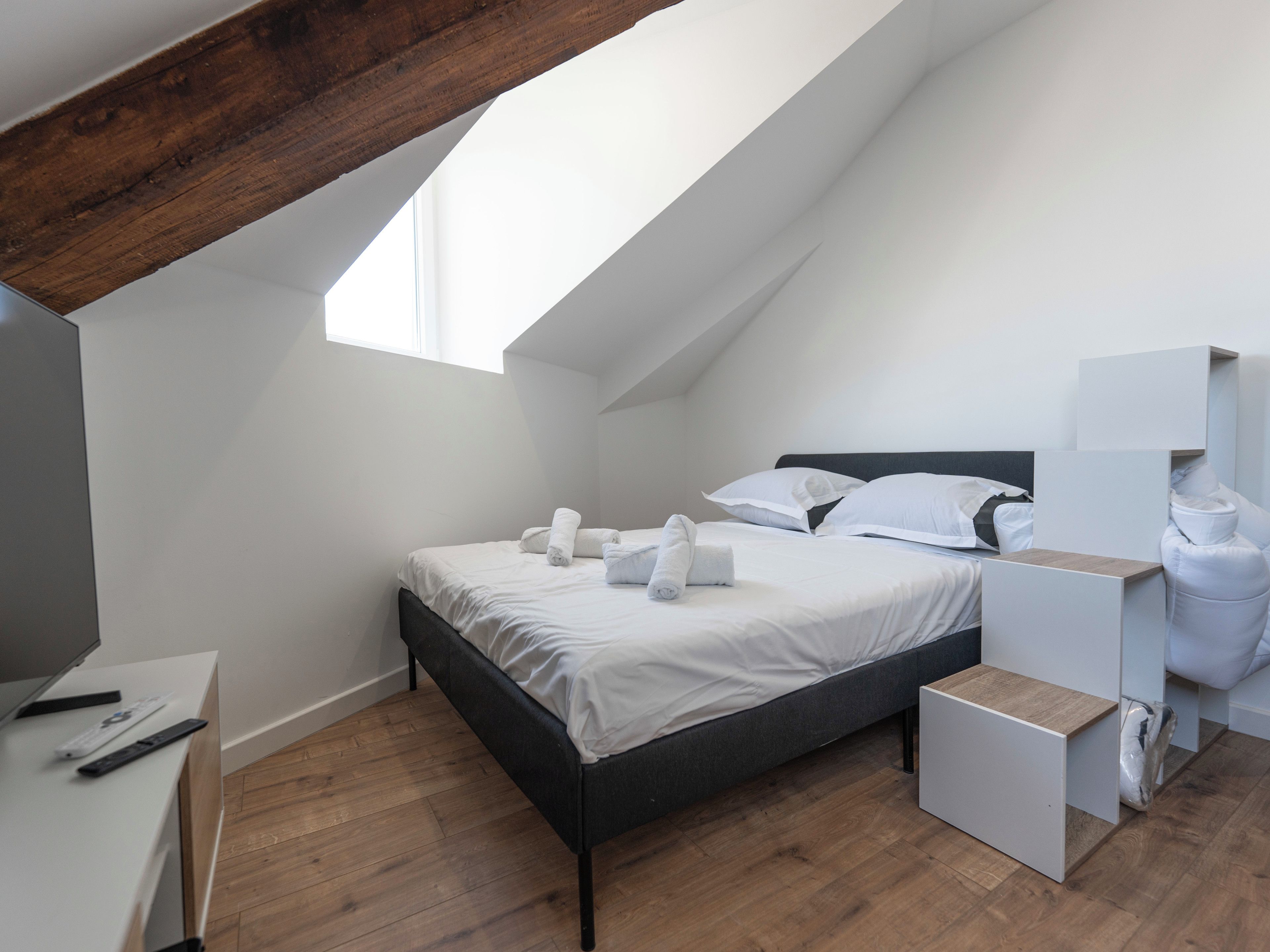 A modern bedroom with a sloped ceiling, a bed with white linens, a wooden desk, and a small bedside table.