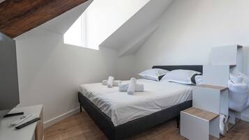 A modern bedroom with a sloped ceiling, a bed with white linens, a wooden desk, and a small bedside table.