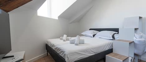 A modern bedroom with a sloped ceiling, a bed with white linens, a wooden desk, and a small bedside table.