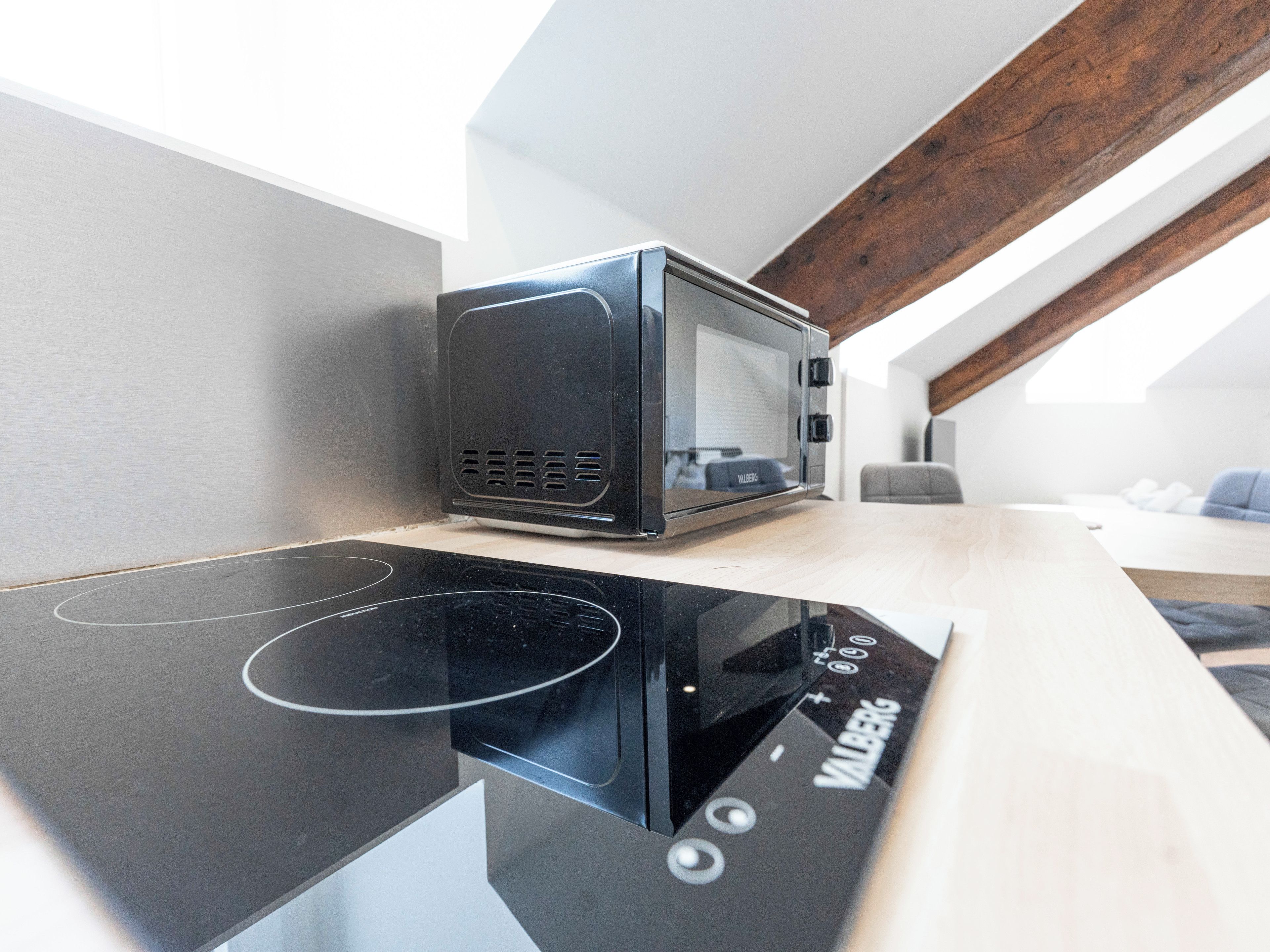 A modern kitchen with a built-in microwave and induction cooktop, a wooden ceiling beam, and a view of a living area.