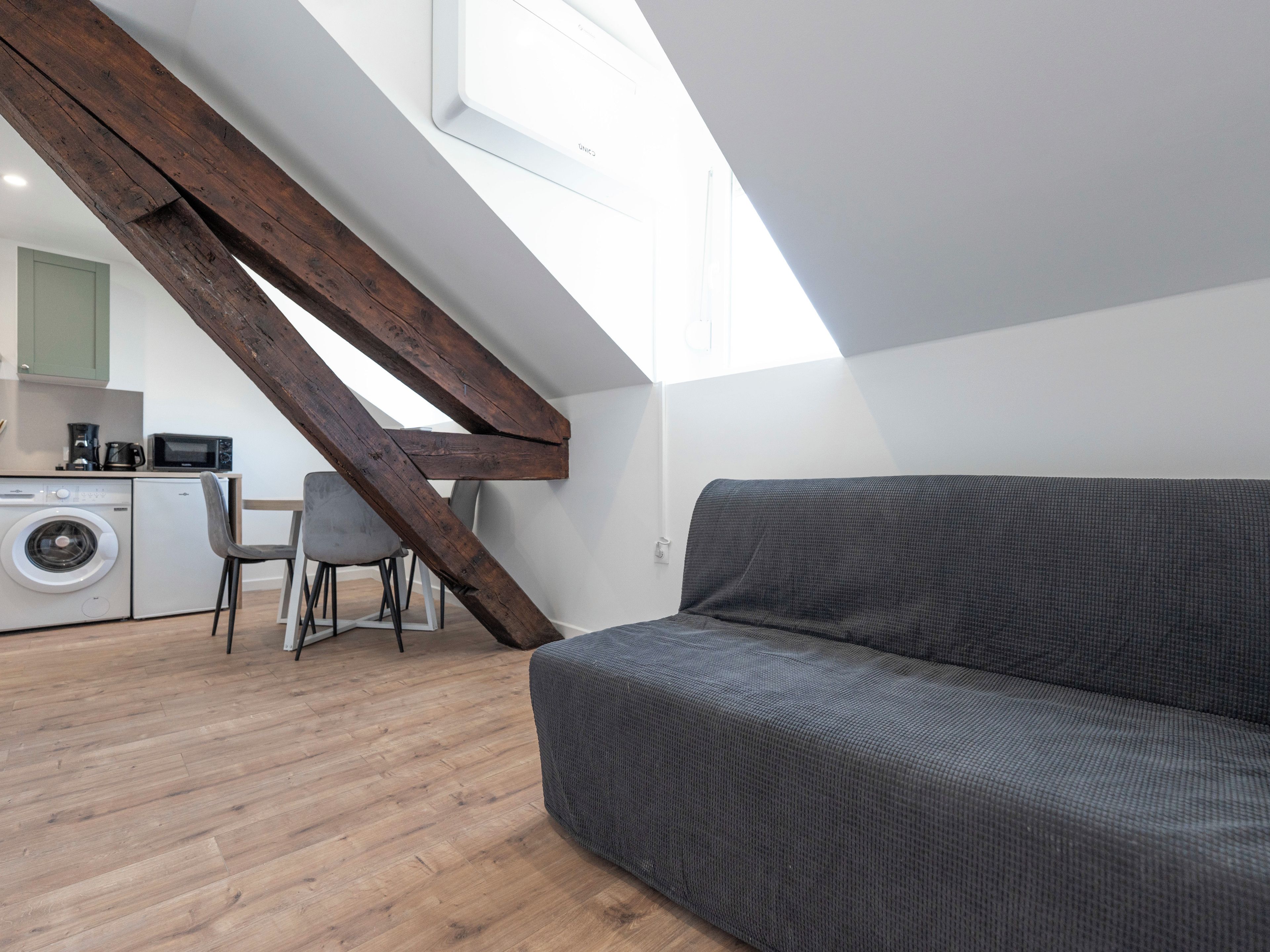 A modern loft bedroom with a sloped ceiling, a grey sofa, a washing machine, and a dining area with a chair and table.