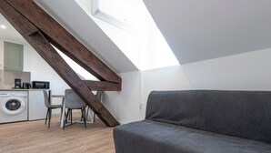Un dormitorio estilo loft moderno con techo inclinado, un sofá gris, una lavadora y una zona de comedor con una silla y mesa.