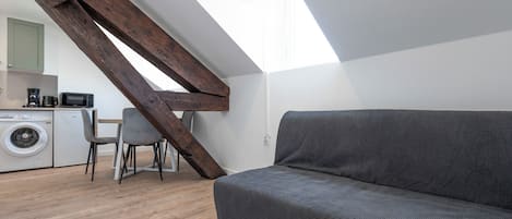 A modern loft bedroom with a sloped ceiling, a grey sofa, a washing machine, and a dining area with a chair and table.