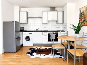 A compact kitchen with white cabinets, a washing machine, a refrigerator, and a dining area with a table and chairs. - Fantastic 1BDR Studio in the Heart of City Centre (Liverpool)