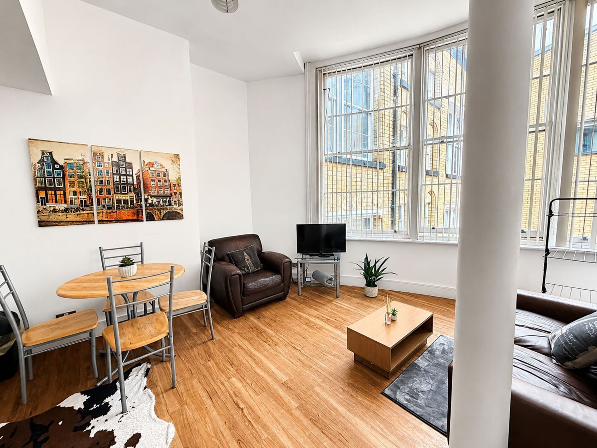 Un soggiorno moderno con pavimento in legno, un divano in pelle marrone, un tavolo da pranzo con sedie, una televisione e un'ampia finestra con vista sugli edifici.