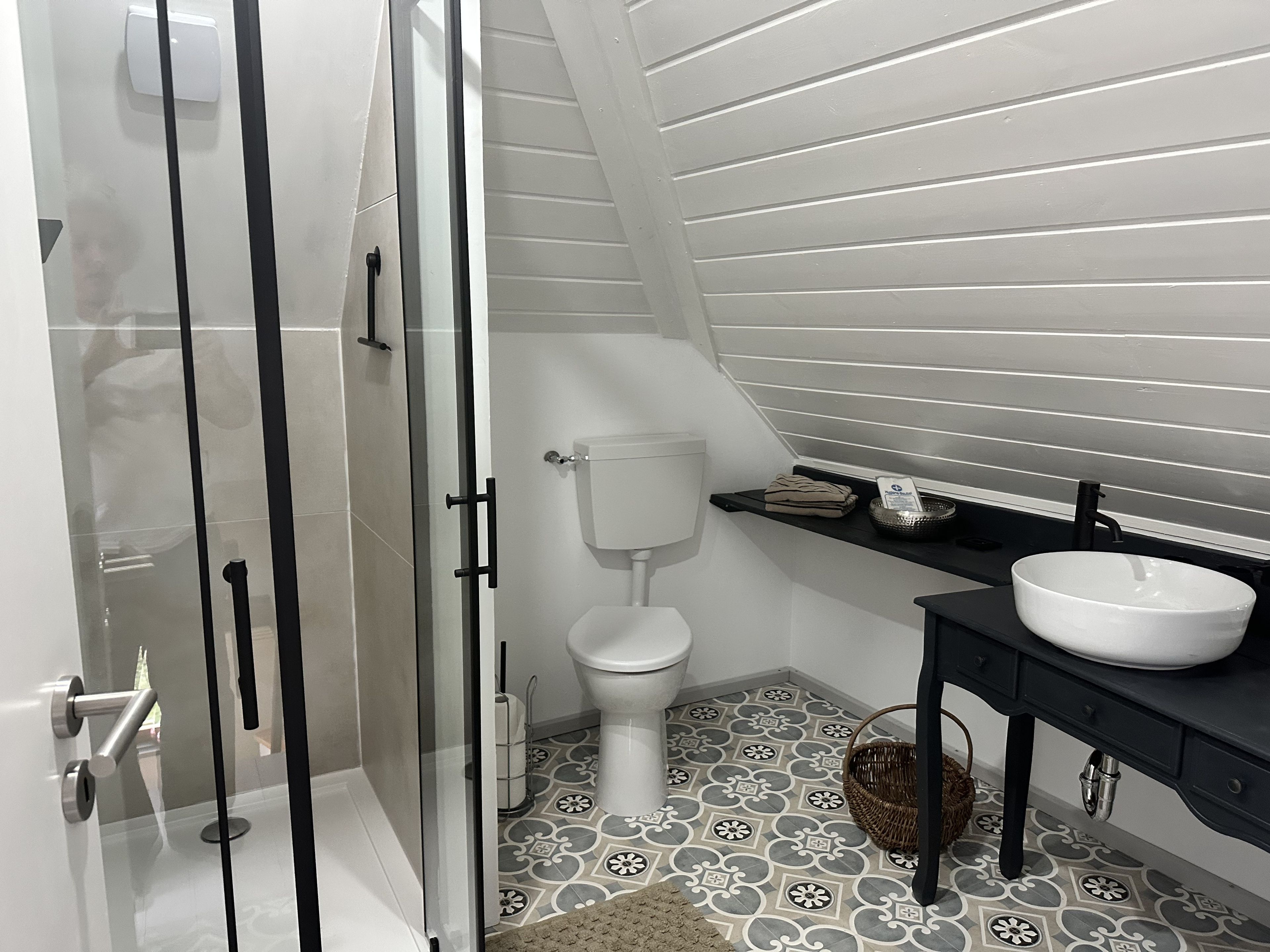 Ein modernes Badezimmer mit Duschkabine aus Glas, einer weißen Toilette, einem schwarzen Waschtisch mit weißem Waschbecken und Fliesenboden mit Muster.