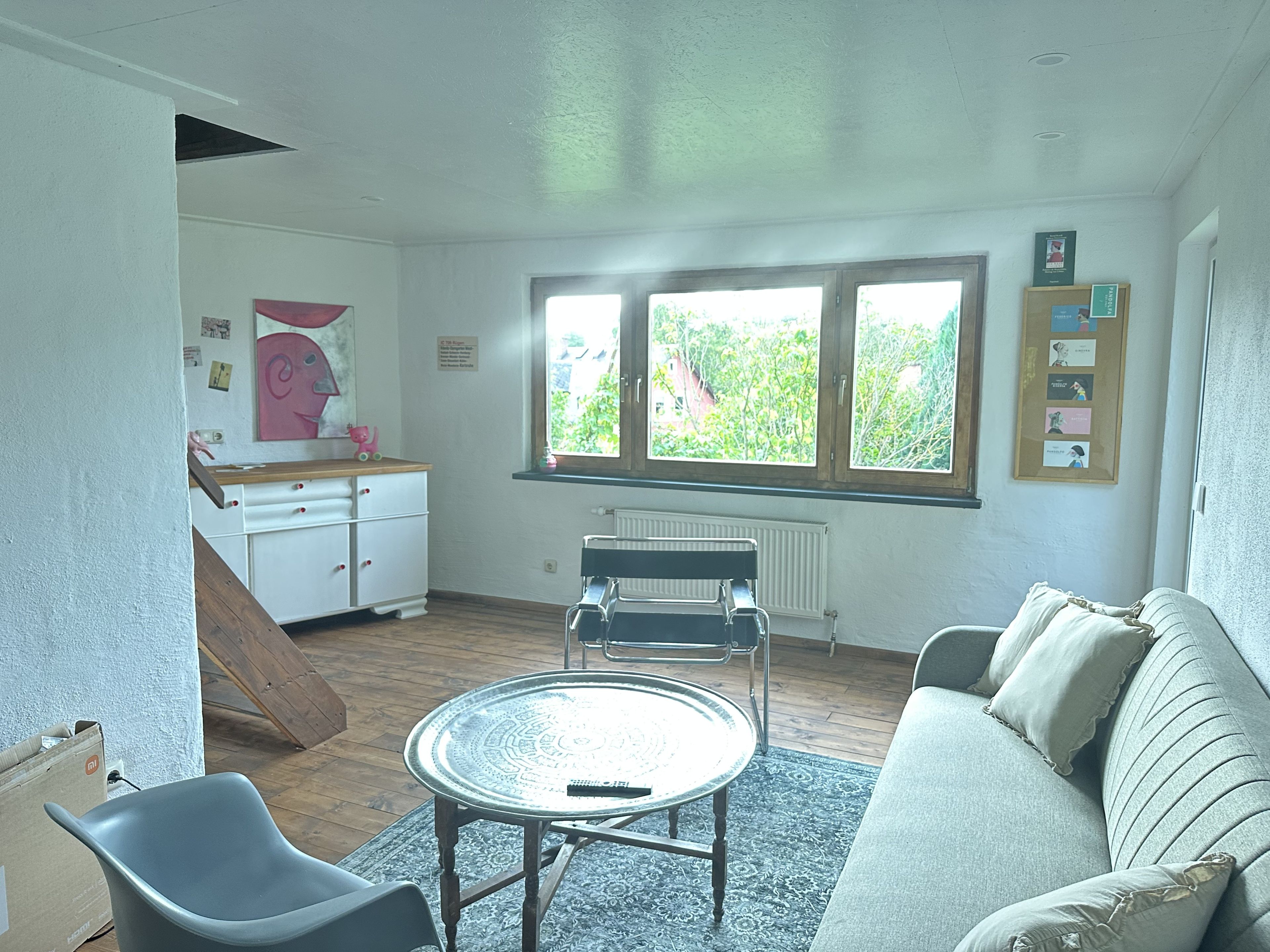 A room with a sofa, a small round table, a chair, a radiator, and a window with a view of greenery.