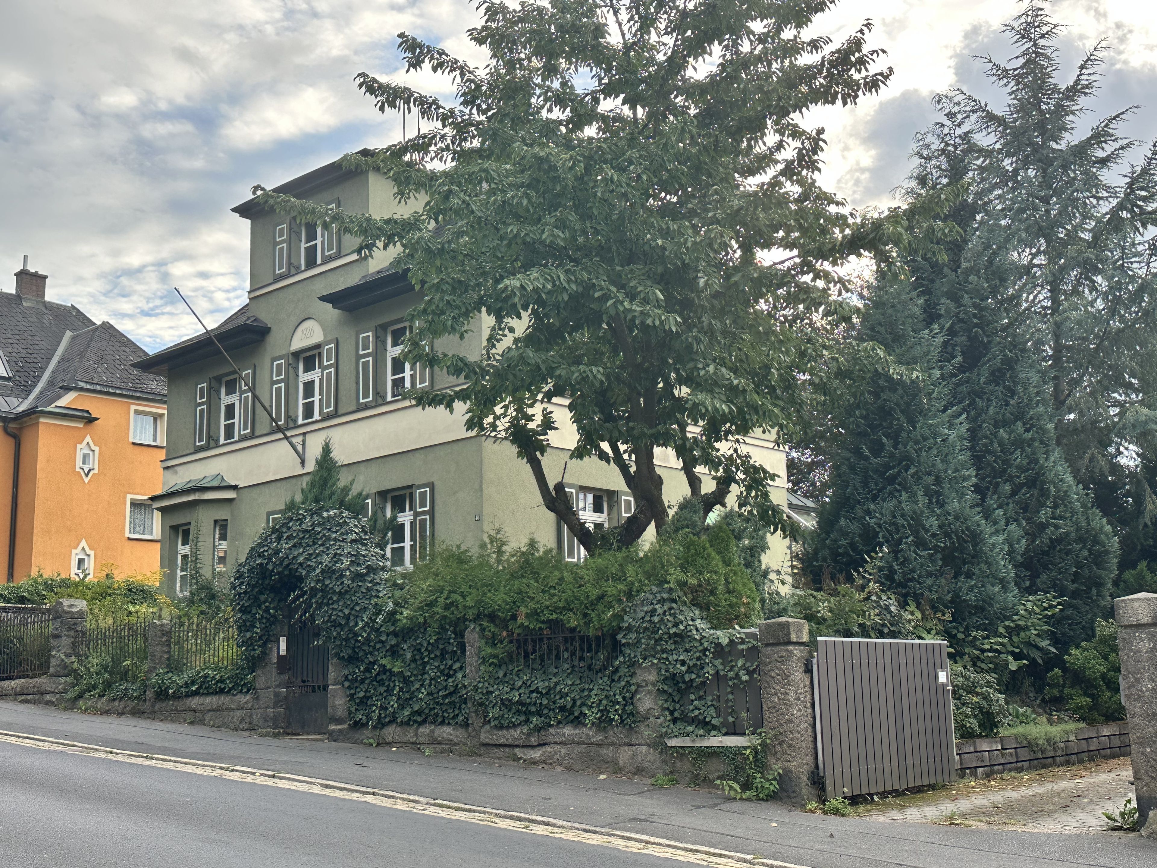Uma casa de dois andares com uma grande árvore em frente, um portão na entrada e uma rua ao fundo.
