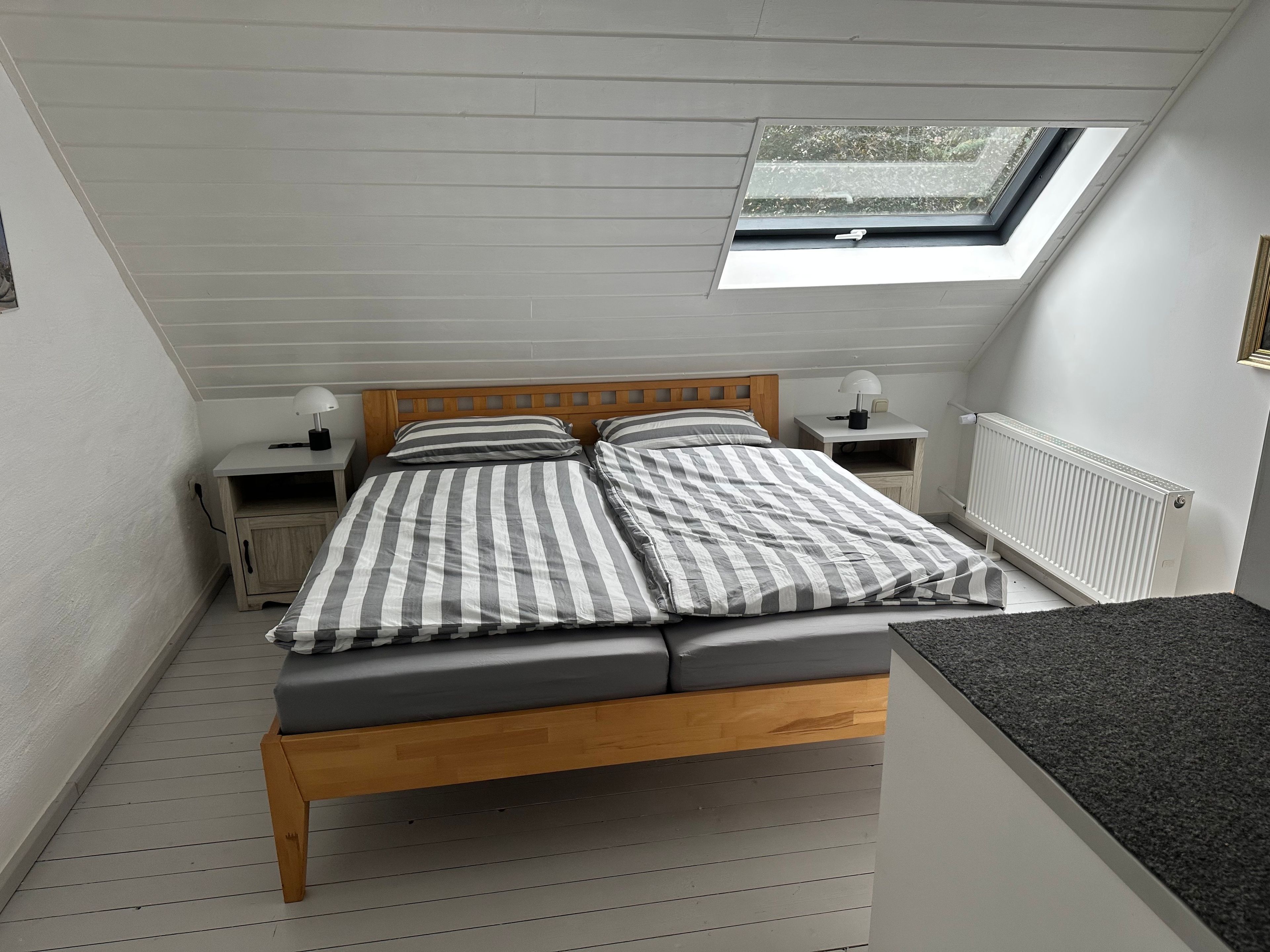 Ein kleines, gemütliches Schlafzimmer mit schrägen Wänden, einem Dachfenster, einem hölzernen Bett, zwei Nachttischen mit Lampen und einem weißen Heizkörper.