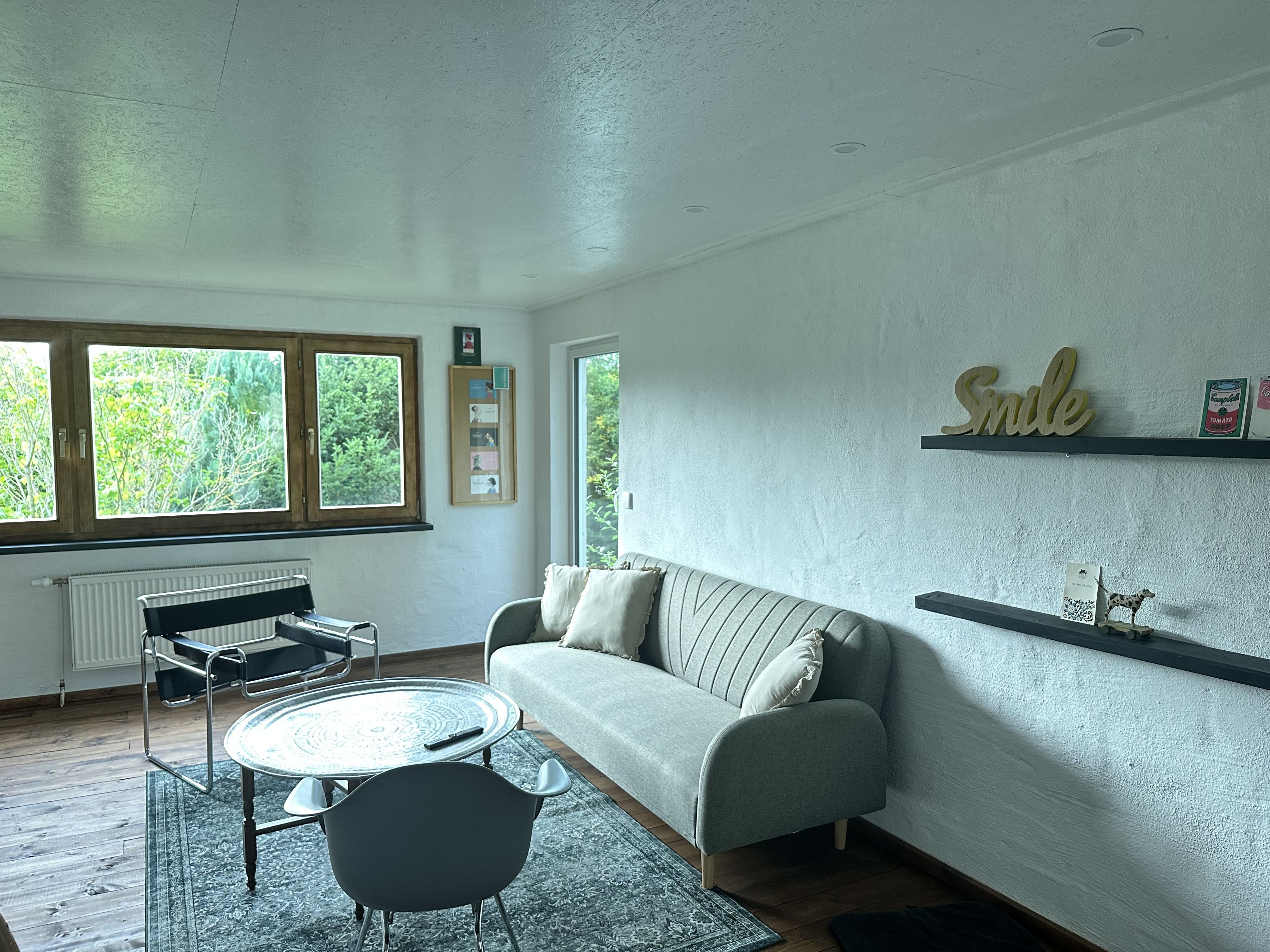 Ein Wohnzimmer mit Sofa, Couchtisch, Sessel und Heizkörper. Fenster bieten Blick auf Grünflächen.
