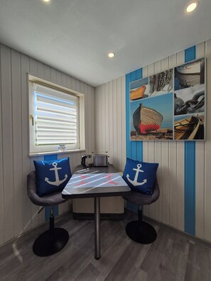 Ein kleiner Essbereich mit einem Tisch und zwei Stühlen, ein Fenster mit Jalousien und eine Wand mit maritimer Dekoration.