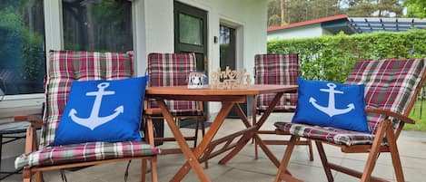 An outdoor seating area with plaid chairs and blue pillows, a wooden table, and a view of a building with large windows.
