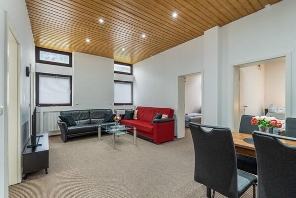 A modern living room with a black leather sofa, a red sofa, and a glass coffee table. There is a dining area with a wooden table and chairs.