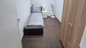 Et lille, aflangt rum med en seng, et sengebord, en lampe, en ventilator og et klædeskab.