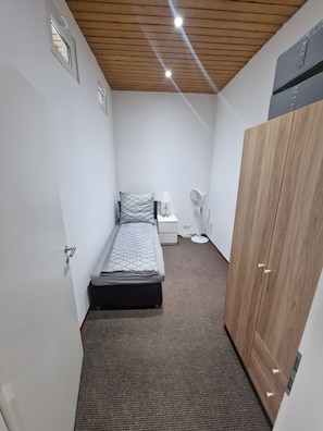 A small, narrow room with a bed, a bedside table, a lamp, a fan, and a wardrobe. - Bright, Modern 3-Room Apartment Near Munich Airport, S-Bahn, 30 Min to City Center (Neufahrn bei Freising)
