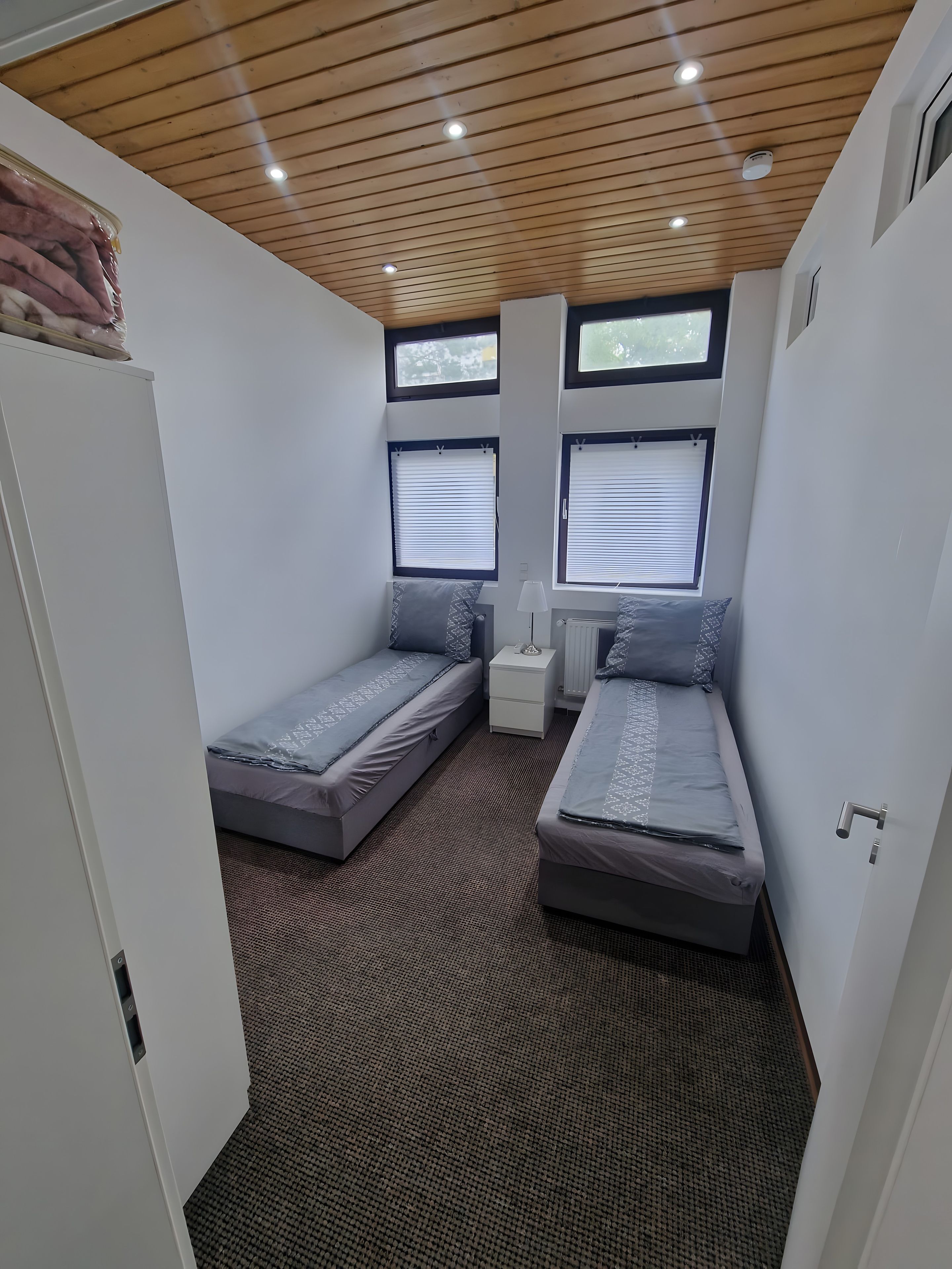 Ein kleines Zimmer mit zwei Einzelbetten, einem kleinen Nachttisch und einem Fenster mit Jalousien.
