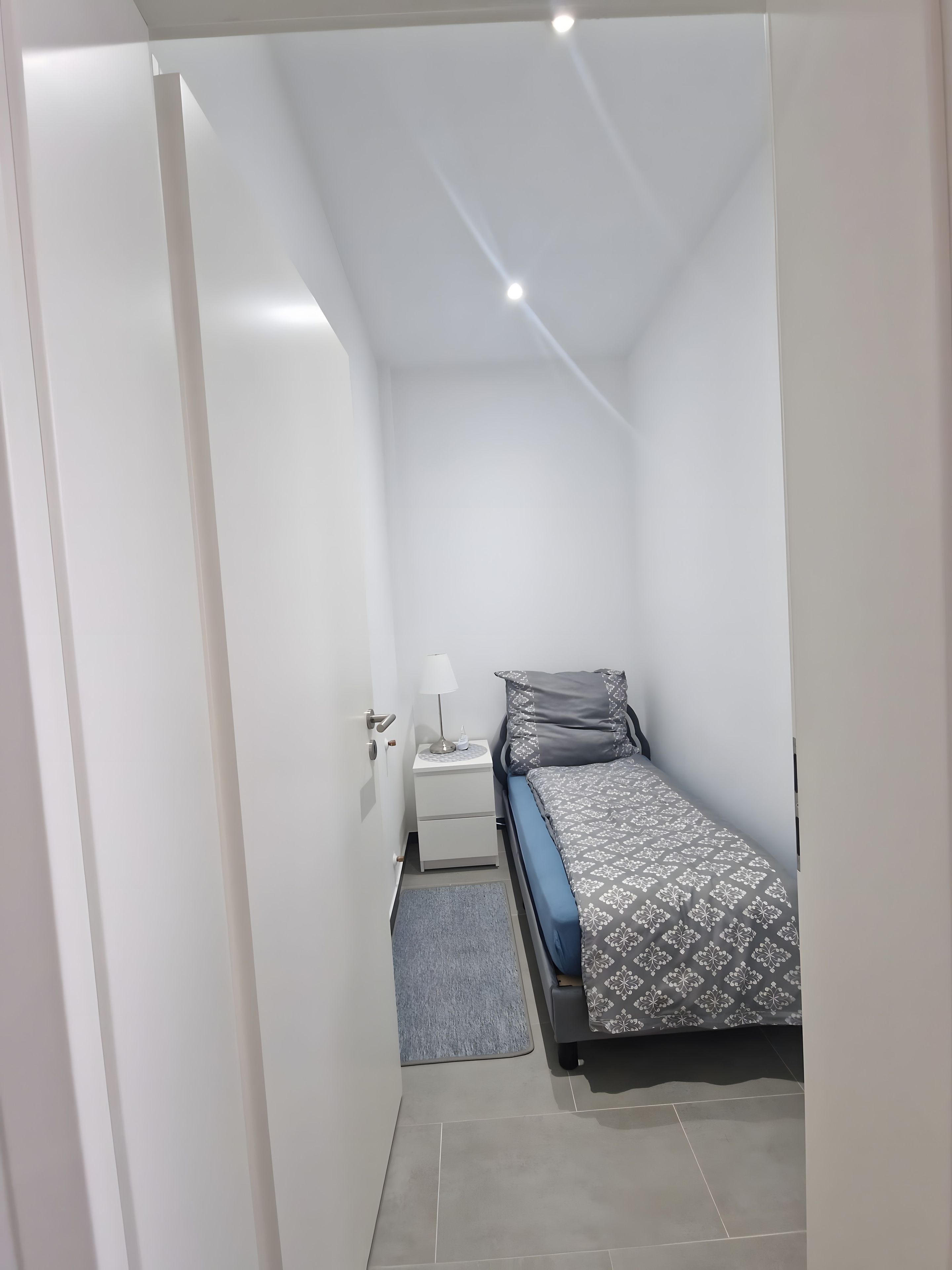 A small, modern bedroom with a single bed, a bedside table with a lamp, and a grey rug.