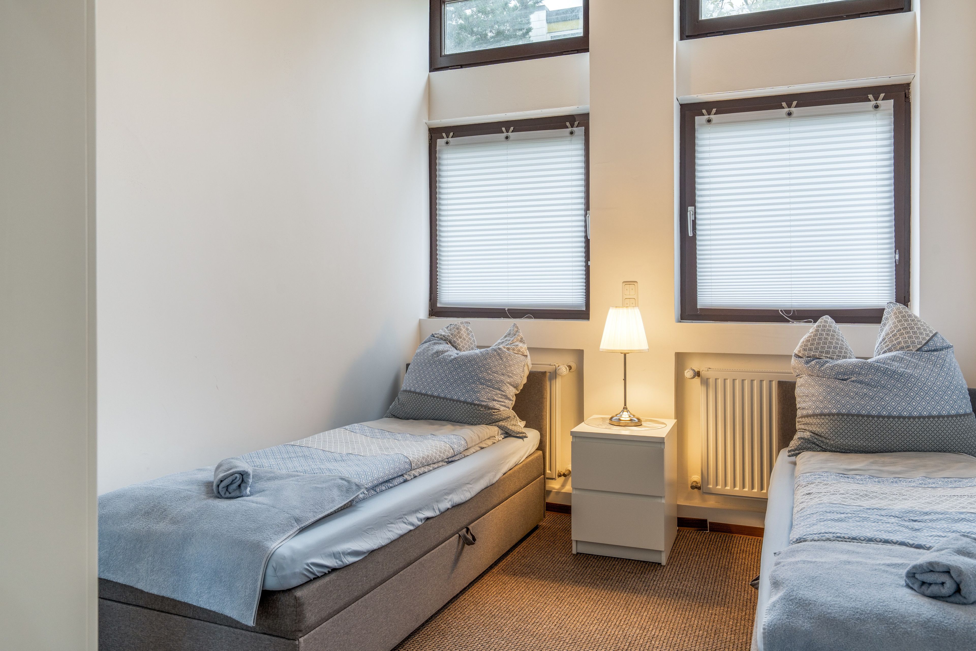 Ein kleines, helles Schlafzimmer mit zwei Einzelbetten, einem Nachttisch mit Lampe und einem Heizkörper.