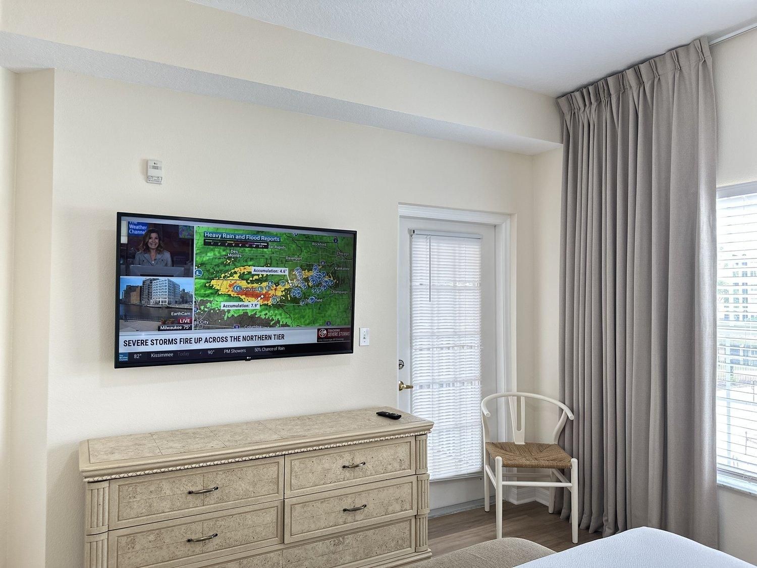 Ein Hotelzimmer mit einem Fernseher, der eine Wettervorhersage zeigt, einer Holzkommode, einem weißen Stuhl und einem Fenster mit Vorhängen.