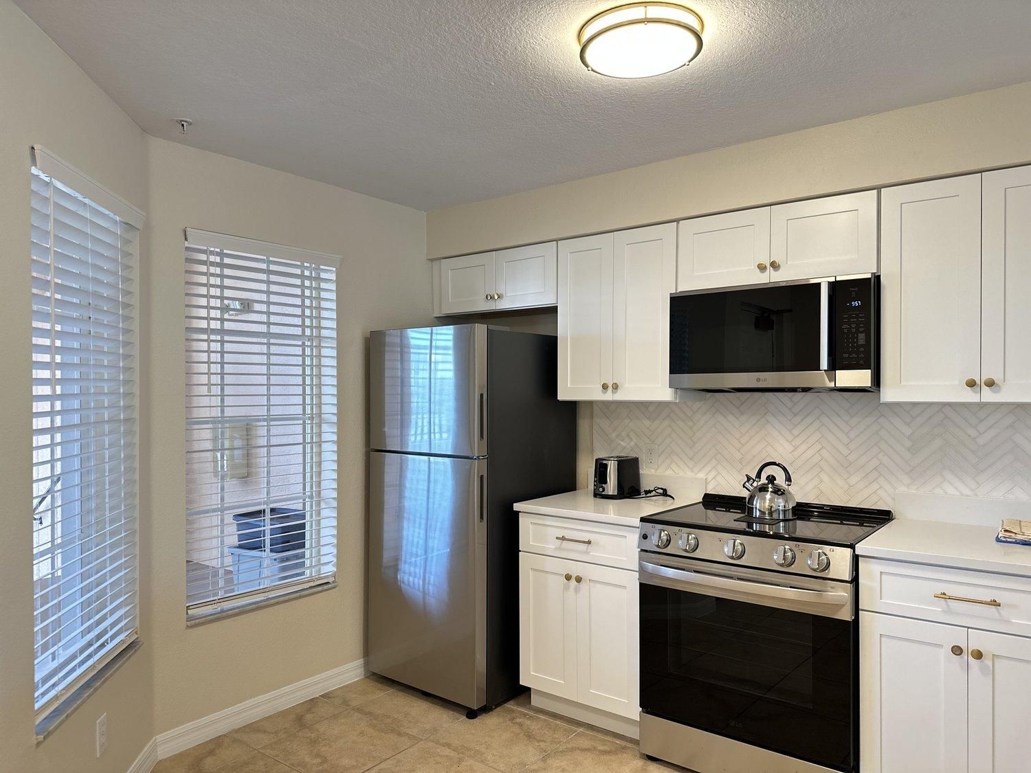A kitchen with white cabinets, a stainless steel refrigerator, a microwave, a stove, and a window with blinds.