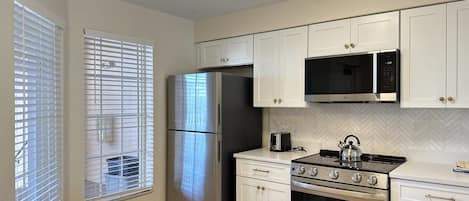 A kitchen with white cabinets, a stainless steel refrigerator, a microwave, a stove, and a window with blinds.