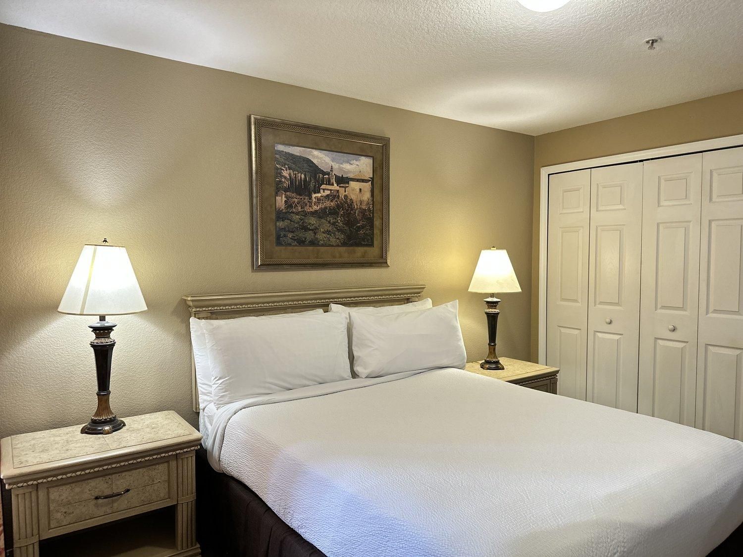 A hotel room with a bed, two bedside lamps, a nightstand, a dresser, a framed picture on the wall, and a set of white doors.