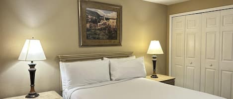 A hotel room with a bed, two bedside lamps, a nightstand, a dresser, a framed picture on the wall, and a set of white doors.
