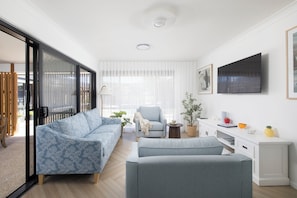 A modern living room with a sofa, armchair, and ottoman, a flat-screen TV, and a window with sheer curtains.