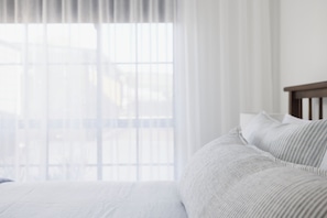A bedroom with a bed, a window with sheer curtains, and a view of buildings outside.