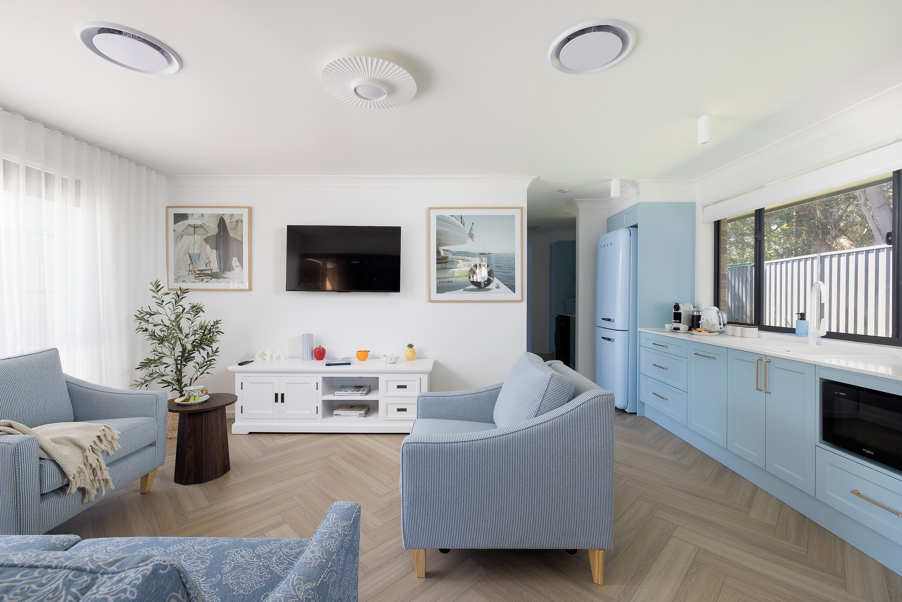 A modern living room with a TV, seating, and a kitchen area with cabinets and a window.