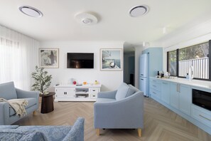 A modern living room with a TV, seating, and a kitchen area with cabinets and a window.