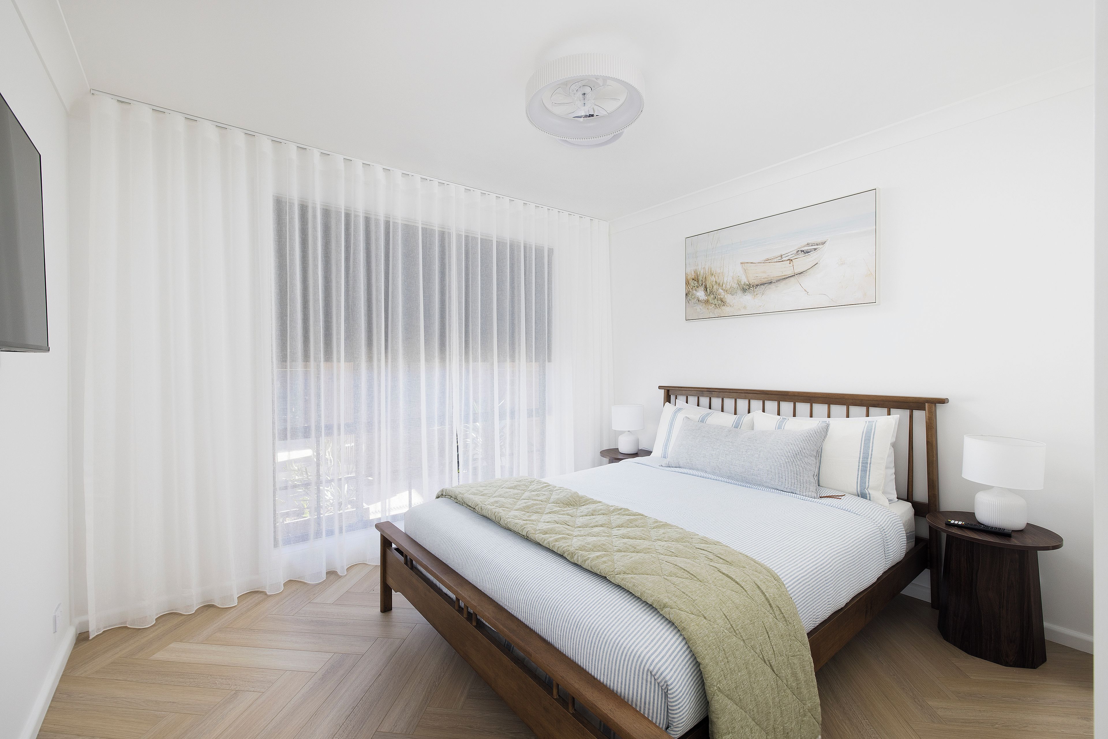A bedroom with a wooden bed, white bedding, a nightstand with a lamp, and a window with sheer curtains.