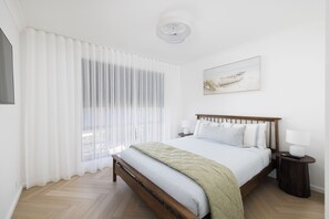 A bedroom with a wooden bed, white bedding, a nightstand with a lamp, and a window with sheer curtains.