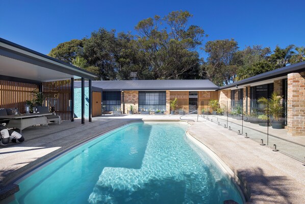 A modern outdoor pool area with a clear blue pool, surrounded by a covered patio with seating and a brick building in the background.