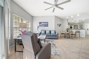 A spacious living area with a grey sofa, a dining table with chairs, a ceiling fan, and artwork on the walls. - 2103 Spacious 2/2 Resort Condo Near Disney (Orlando)